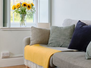 A sofa with cushions and a yellow throw next to a window with sunflowers in a vase at Cove Cottage in Porthgwarra