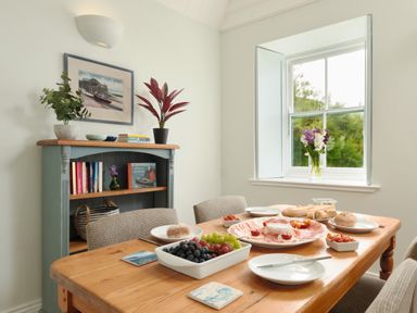 A dining area with a wooden table set with plates bread and fruit beside a window and a bookshelf with books and plants at Cove Cottage in Porthgwarra