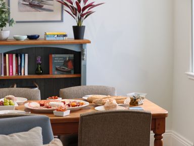 A dining table with bread, grapes, olives, and a charcuterie plate in a room with a bookshelf at Cove Cottage in Porthgwarra