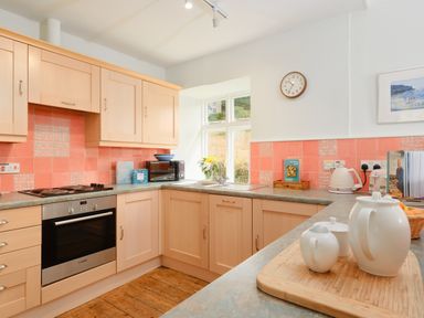 A kitchen with wooden cabinets pink tiled backsplash an oven a microwave and a teapot set on the counter at Cove Cottage in Porthgwarra