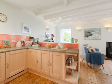 A kitchen with light wood cabinets and coral tile backsplash next to a living area with blue chairs and a fireplace at Cove Cottage in Porthgwarra