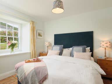 A bedroom with a large bed with blue headboard and patterned pillows two wooden bedside tables with lamps and a window with yellow curtains and flowers at Cove Cottage in Porthgwarra