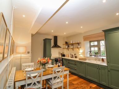 A kitchen with green cabinets a wooden dining table set with plates glasses and flowers at Omas Cottage in Moreton In Marsh