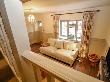 A living room with a beige sofa under a window with floral curtains and a floor lamp at Oma's Cottage in Moreton-In-Marsh