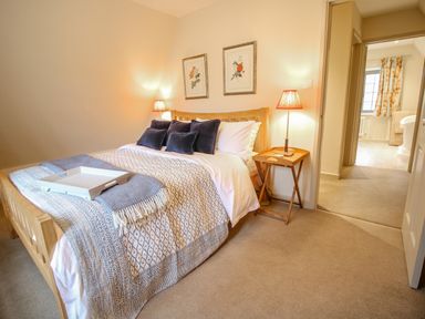 A bedroom with a wooden bed frame and patterned bedding with cushions and bedside lamps at Omas Cottage in Moreton-In-Marsh