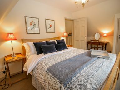 A bedroom with a wooden bed with patterned bedding and dark pillows two bedside tables with lamps a desk with a mirror and chair Oma's Cottage in Moreton-In-Marsh