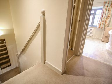 A carpeted staircase with a handrail and a hallway leading to a bathroom with a window and floral curtains at Omas Cottage in Moreton In Marsh