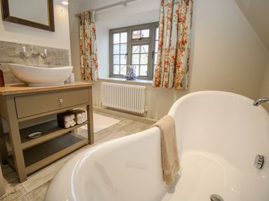 A bathroom with a freestanding bathtub and a vanity unit under a window with floral curtains at Omas Cottage in Moreton-In-Marsh