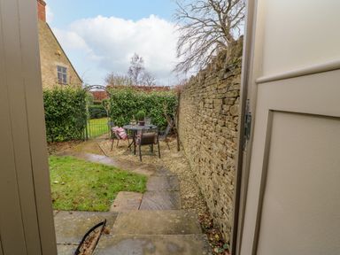 A small garden with stone steps leading to a seating area with chairs and a table surrounded by a stone wall and greenery at Omas Cottage in Moreton In Marsh