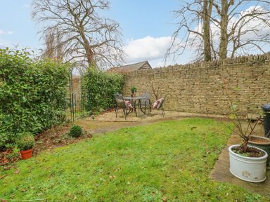 A garden with a stone wall hedge a seating area with table and chairs and potted plants at Omas Cottage in Moreton-In-Marsh
