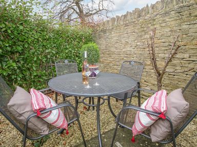An outdoor patio with a round metal table and four chairs with cushions against a stone wall at Omas Cottage in Moreton-In-Marsh