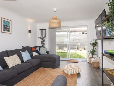 A living room with a dark sectional sofa cushions a striped floor pillow and a sliding glass door overlooking a garden at Dolwyn in Llandegfan