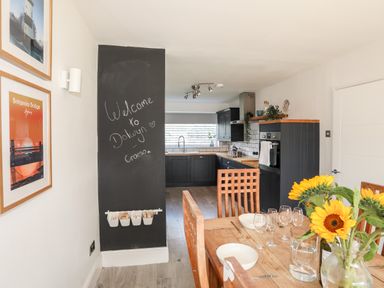A dining area with a wooden table set with bowls and wine glasses next to a kitchen with dark cabinets and a window at Dolwyn in Llandegfan