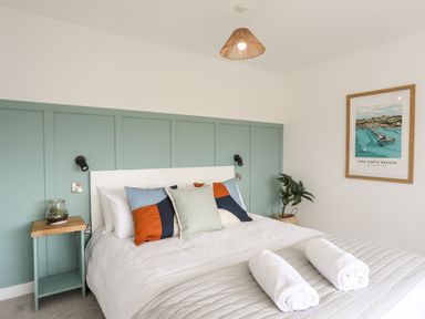 A bedroom with a bed with pillows and rolled towels bedside table with glass jar plant on floor and framed picture on wall at Dolwyn in Llandegfan