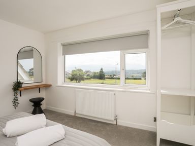 A bedroom with a large window a wall-mounted mirror a small black stool and an open closet at Dolwyn in Llandegfan
