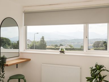A corner with a window and a wooden shelf with a mirror a green stool and plants at Dolwyn in Llandegfan