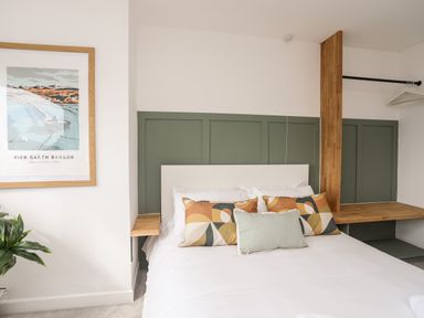 A bedroom with a bed with multiple pillows a framed picture on the wall and wooden shelves at Dolwyn in Llandegfan