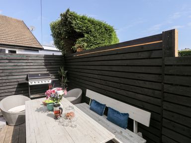 An outdoor patio area with a white wooden table and bench with cushions two chairs a barbecue grill and a flower vase at Dolwyn in Llandegfan