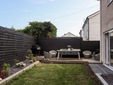 A backyard with a lawn a wooden deck with a table chairs and a barbecue grill at Dolwyn in Llandegfan