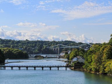 A river with bridges and trees on either side and a suspension bridge in the background at Dolwyn in Llandegfan