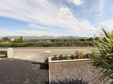 A view of a road with a stone wall and a flower bed with plants with hills in the background at Dolwyn in Llandegfan