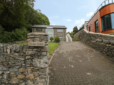 A driveway leading to a stone house with a lawn and a wooden sign saying Tan Twr next to a modern building at Tan Twr in Llanfairpwll