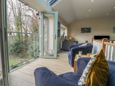 A seating area with blue armchairs and a couch near an open glass door overlooking trees at Tan Twr in Llanfairpwll