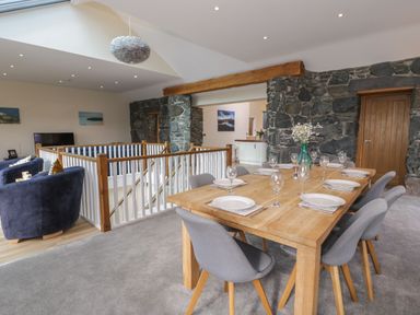 A dining area with a wooden table set with plates and glasses and grey chairs in a room with stone walls and a sitting area at Tan Twr in Llanfairpwll