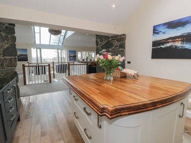 A kitchen island with a wooden countertop and flowers in a vase overlooking a living area with stone walls and large windows at Tan Twr in Llanfairpwll