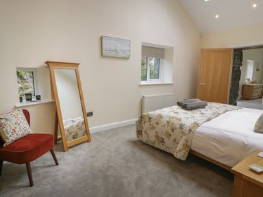 A bedroom with a bed covered with a floral blanket a wooden mirror leaning against the wall a red chair with a floral pillow and a wooden door at Tan Twr in Llanfairpwll