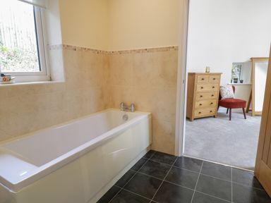 A bathroom with a bathtub next to a window and a door leading to a room with a wooden dresser and a red chair at Tan Twr in Llanfairpwll