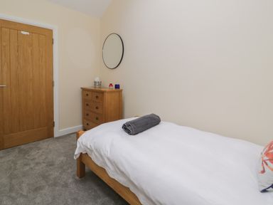 A bedroom with a single wooden bed with white bedding and a rolled gray towel on top a wooden chest of drawers with items on it and a round mirror on the wall at Tan Twr in Llanfairpwll