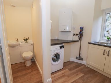 A utility room with washing machine and vacuum cleaner next to a small bathroom with toilet and sink at Tan Twr in Llanfairpwll