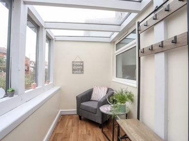 A small sunroom with a grey armchair a small table with a plant a coat rack and windows at Beeches Bach in Prestatyn