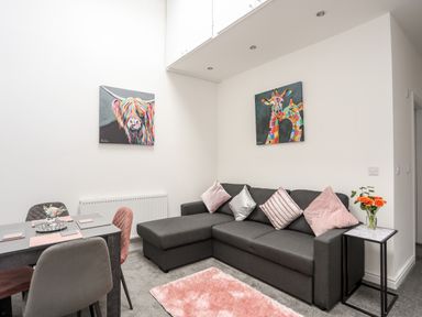 A living room with a gray sectional sofa and pink cushions next to a dining table with four chairs and colorful animal paintings on the walls at Beeches Bach in Prestatyn
