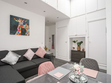 A living room with a dark sofa and pink and silver cushions a colorful giraffe painting plants and a dining table with flowers at Beeches Bach in Prestatyn