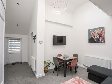 A dining area with a black table and four chairs below a wall-mounted television and colorful animal painting at Beeches Bach in Prestatyn