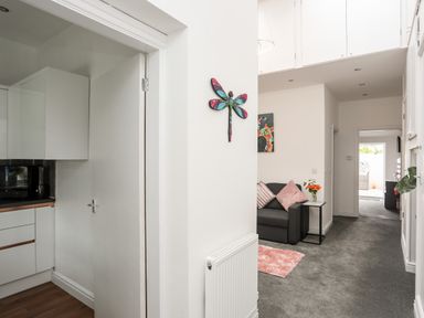 A hallway with a gray sofa and pink cushions next to a small table with flowers and a kitchen on the left at Beeches Bach in Prestatyn