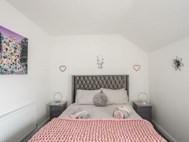 A bedroom with a double bed gray headboard pink blanket bedside tables with lamps and wall decorations at Beeches Bach in Prestatyn