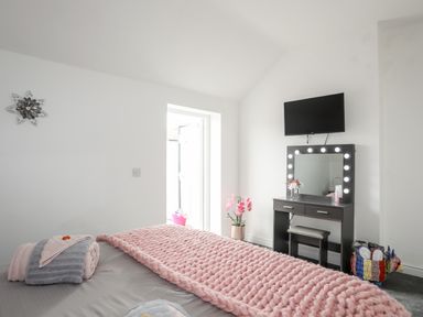 A bedroom with a bed covered in a pink blanket and towels with a dressing table with lights and a TV mounted on the wall at Beeches Bach in Prestatyn