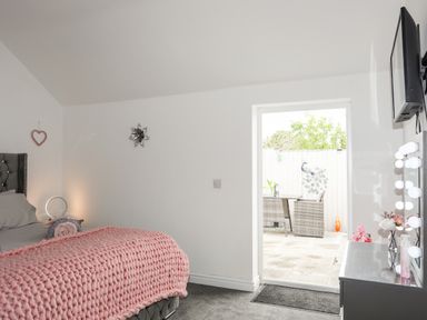 A bedroom with a bed covered by a pink knitted blanket and an open door leading to an outdoor seating area at Beeches Bach in Prestatyn