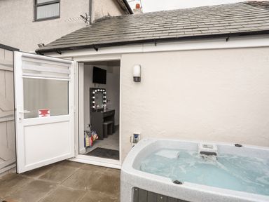 A hot tub on a patio outside a building with an open door showing a vanity table and television inside at Beeches Bach in Prestatyn