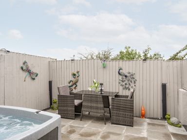 A patio area with a hot tub wicker chairs and a glass table against a fenced wall with garden decorations at Beeches Bach in Prestatyn