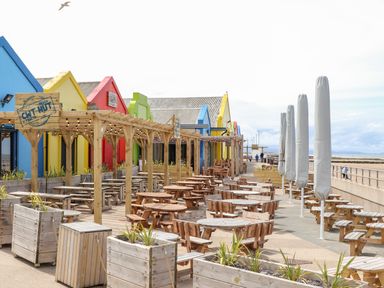 An outdoor seating area with wooden tables and benches by colorful buildings and a beachside promenade at Beeches Bach in Prestatyn
