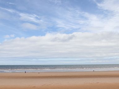 A sandy beach with two people walking near the water and wind turbines visible on the horizon at Beeches Bach in Prestatyn