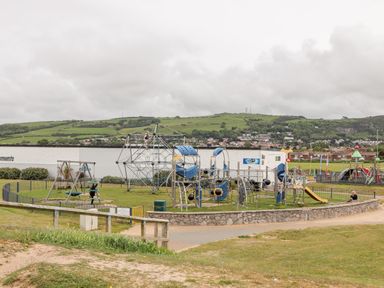 An outdoor playground with climbing structures swings and a slide surrounded by a fence with hills and houses in the background at Beeches Bach in Prestatyn