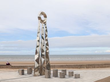 A metallic sculpture with three tall irregular pillars and a circular top near the beach with a person sitting on a bench at Beeches Bach in Prestatyn