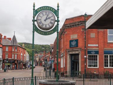 A street scene with a large clock reading prestatyn 1896-1996 in the foreground and red brick buildings along the road at Beeches Bach in Prestatyn