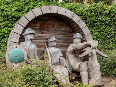 A wooden sculpture of three medieval warriors with helmets and shields surrounded by greenery