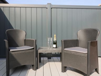 Two wicker chairs with cushions and a small table with a bottle and two glasses on a deck with a grey fence at Chalet 5 in Sherburn